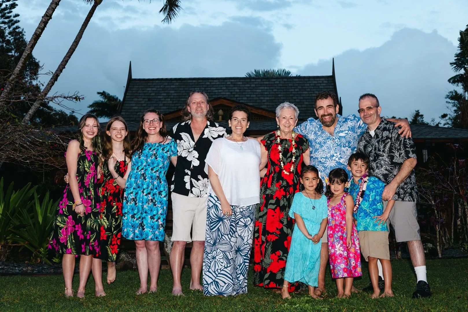 A group of ten people, including children and adults, dressed in tropical and floral clothing, standing outdoors on grass in front of a traditional-style building with a thatched roof, during early evening or dusk.