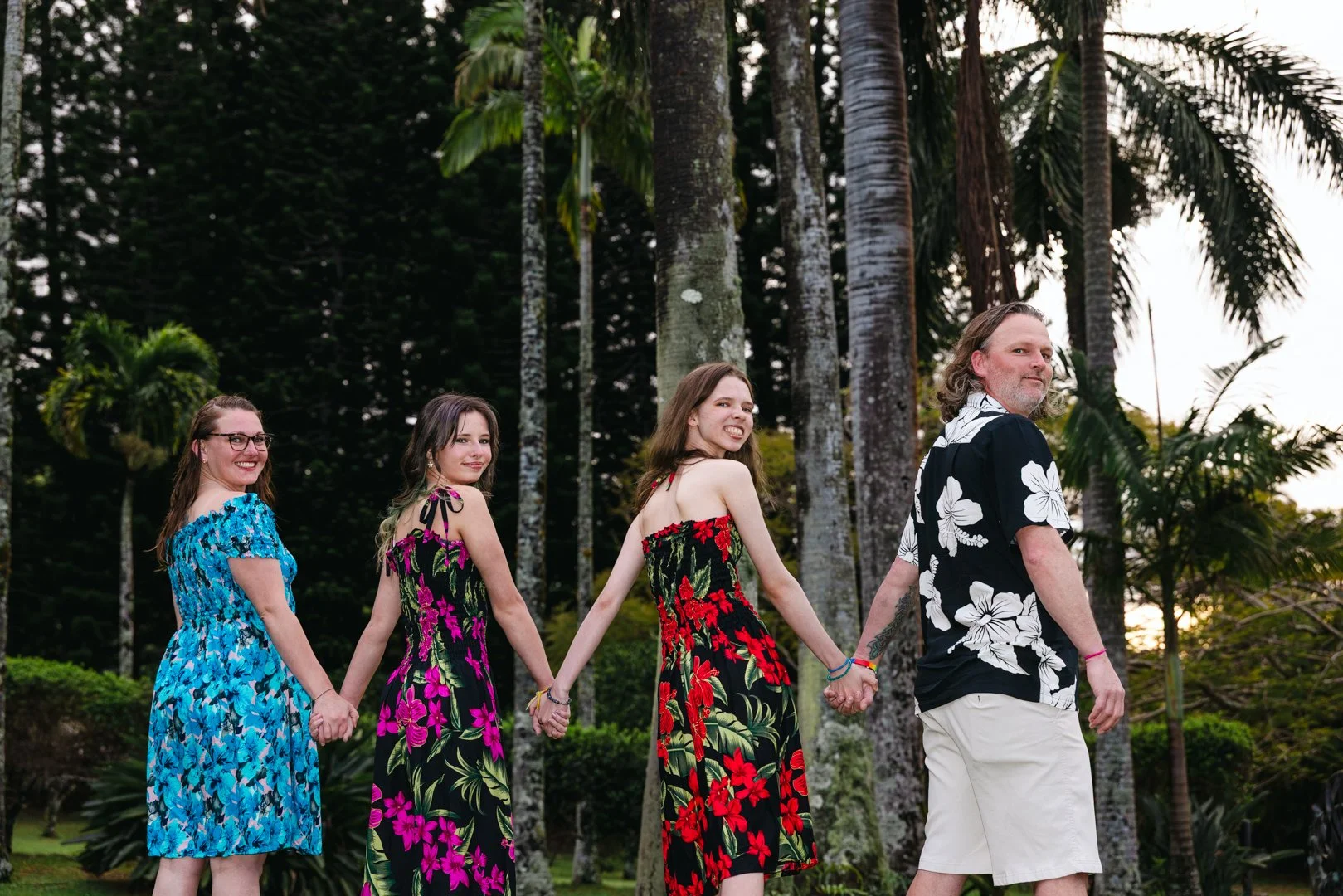 Four people, three women and one man, holding hands and walking in a tropical outdoor setting with tall palm trees in the background. The women are wearing colorful floral dresses, and the man is wearing a black shirt with white flowers and light-col