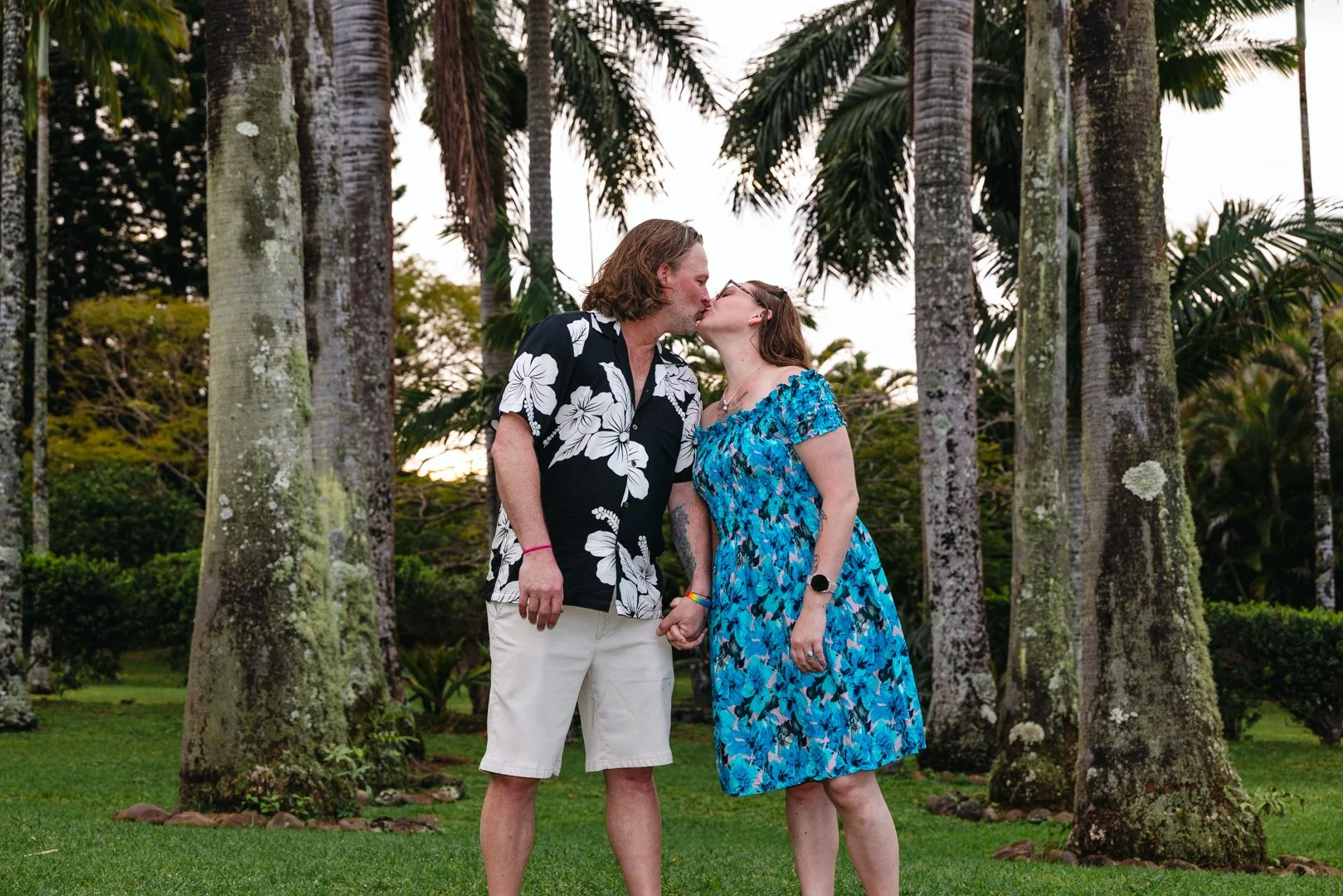 A couple in Hawaiian shirts and floral dresses sharing a kiss and holding hands in a lush, tropical park with tall palm trees.