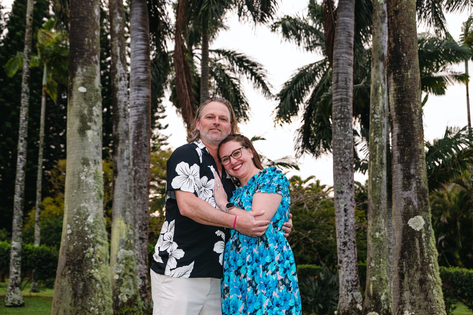 A man and a woman are hugging outdoors among tall trees with a lush background.