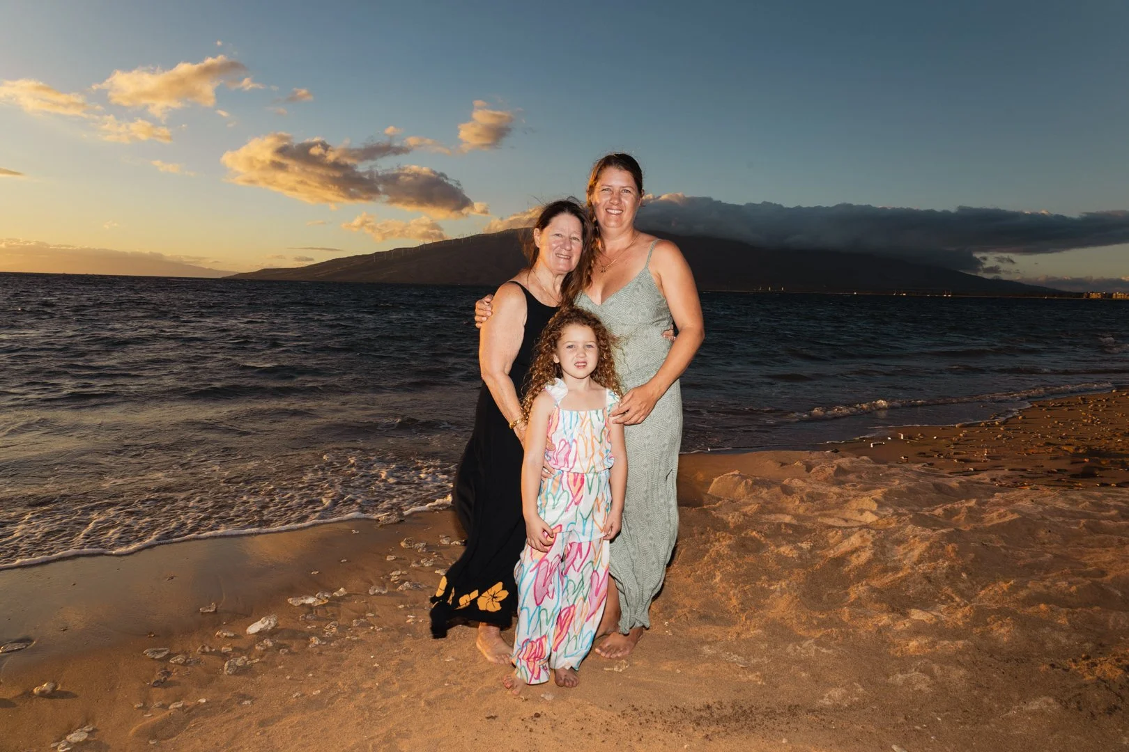 Three women and a girl standing on a sandy beach at sunset with a mountain and ocean in the background.