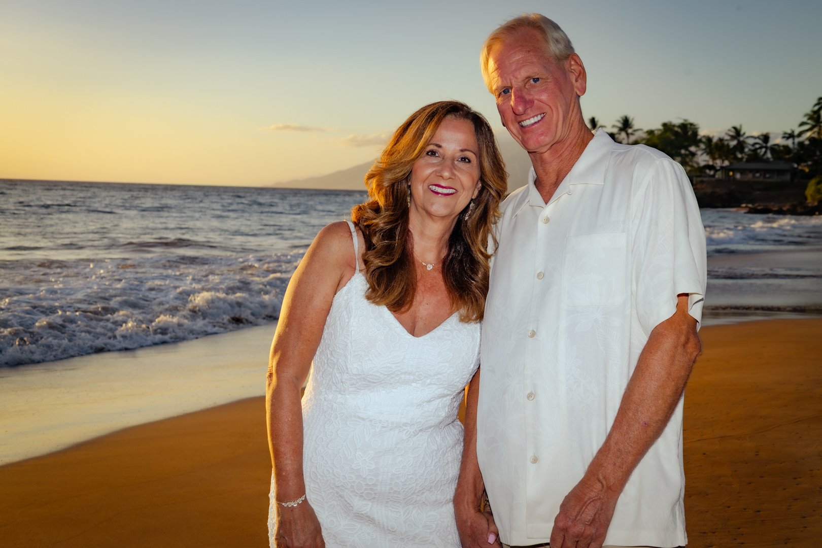 A smiling older couple stands on a beach at sunset. The woman wears a white dress and the man wears a white short-sleeved shirt. The ocean and palm trees are in the background.