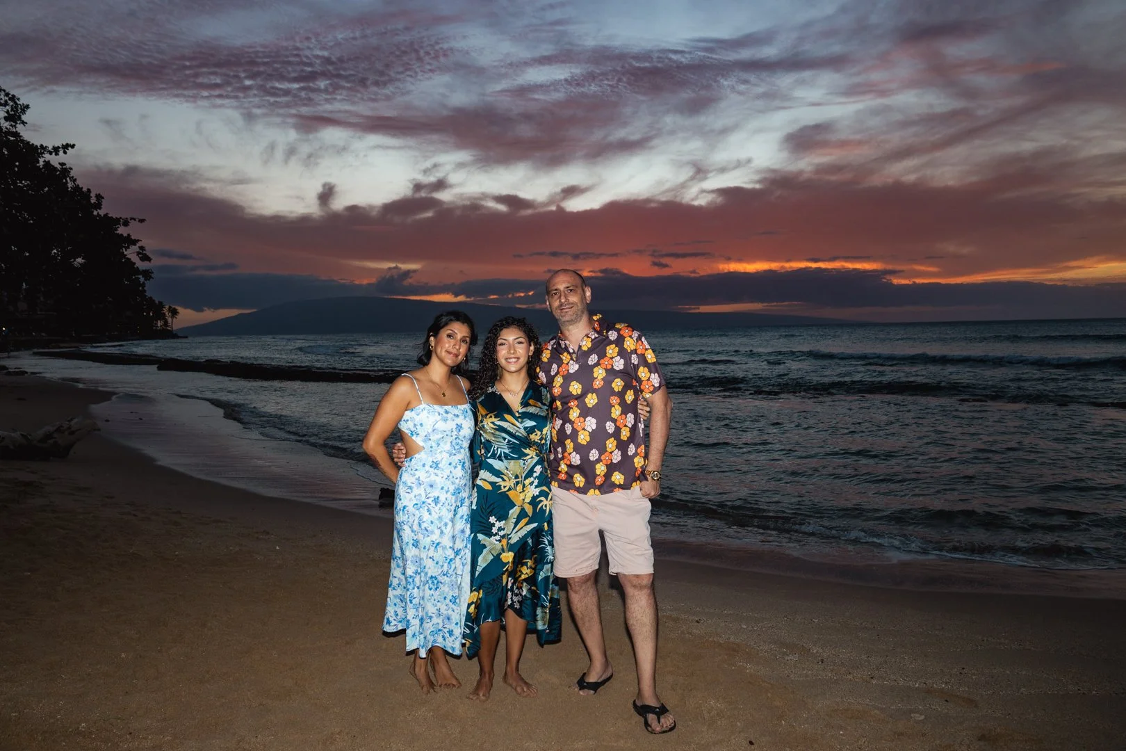 A group of three people standing on a beach during sunset, with two women and one man dressed in summer clothing, smiling at the camera.
