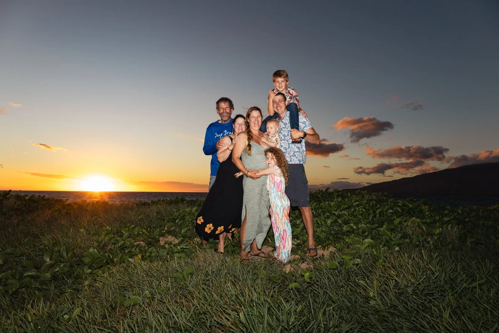 A family of seven standing outdoors during sunset. The family members are smiling and standing on grass with a sunset in the background.