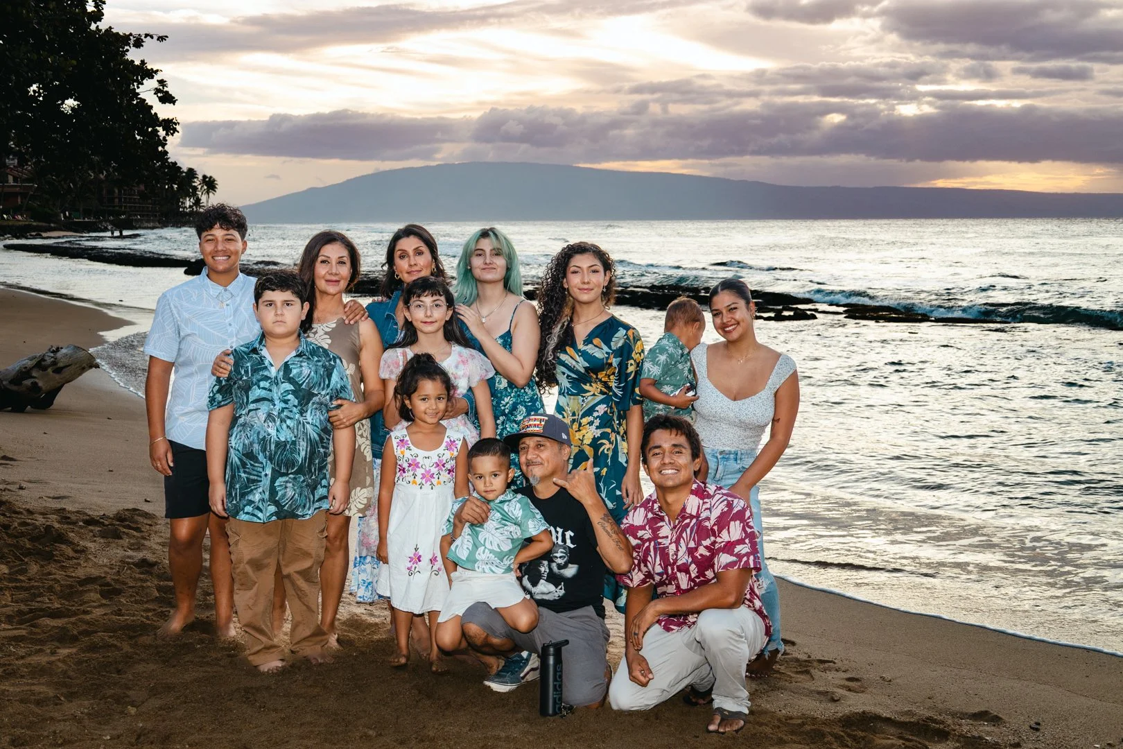 A multi-generational family group photo on a beach during sunset, with ocean waves, rocks, and a mountain in the background. The group includes adults and children dressed in casual, colorful tropical clothing.