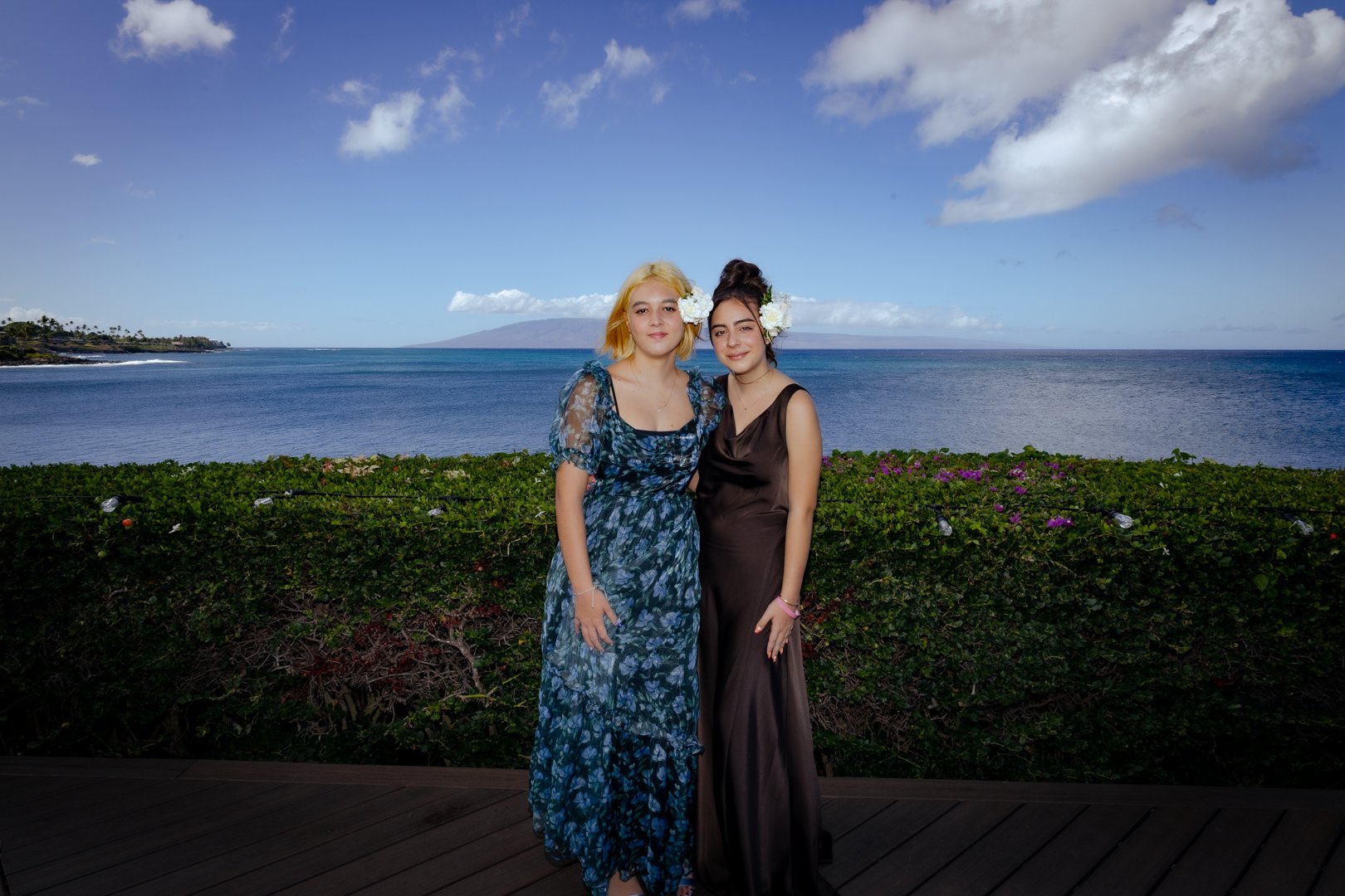 Two women standing outdoors by the ocean, with a landscape of water and clouds in the background. One woman is wearing a blue floral dress, and the other is in a black dress. Both women are wearing flowers in their hair and are smiling.