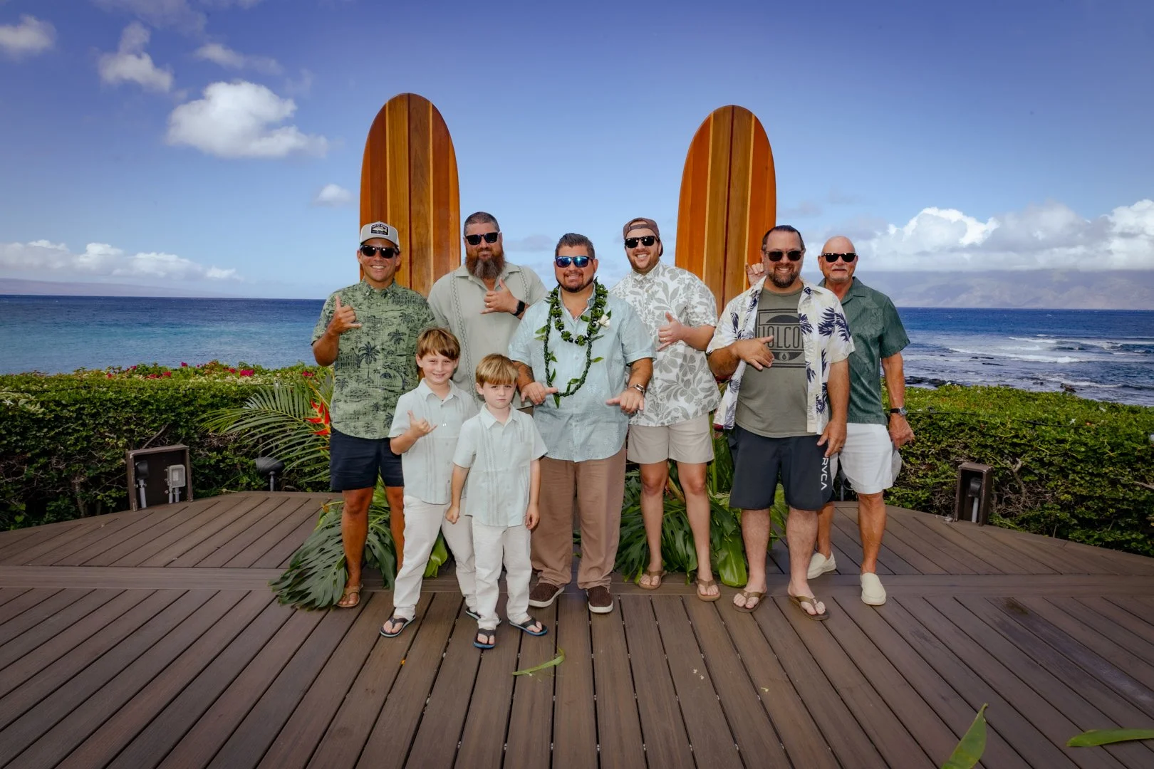 Group of men and boys on a wooden deck at the beach, with surfboards behind them, celebrating during a tropical vacation.