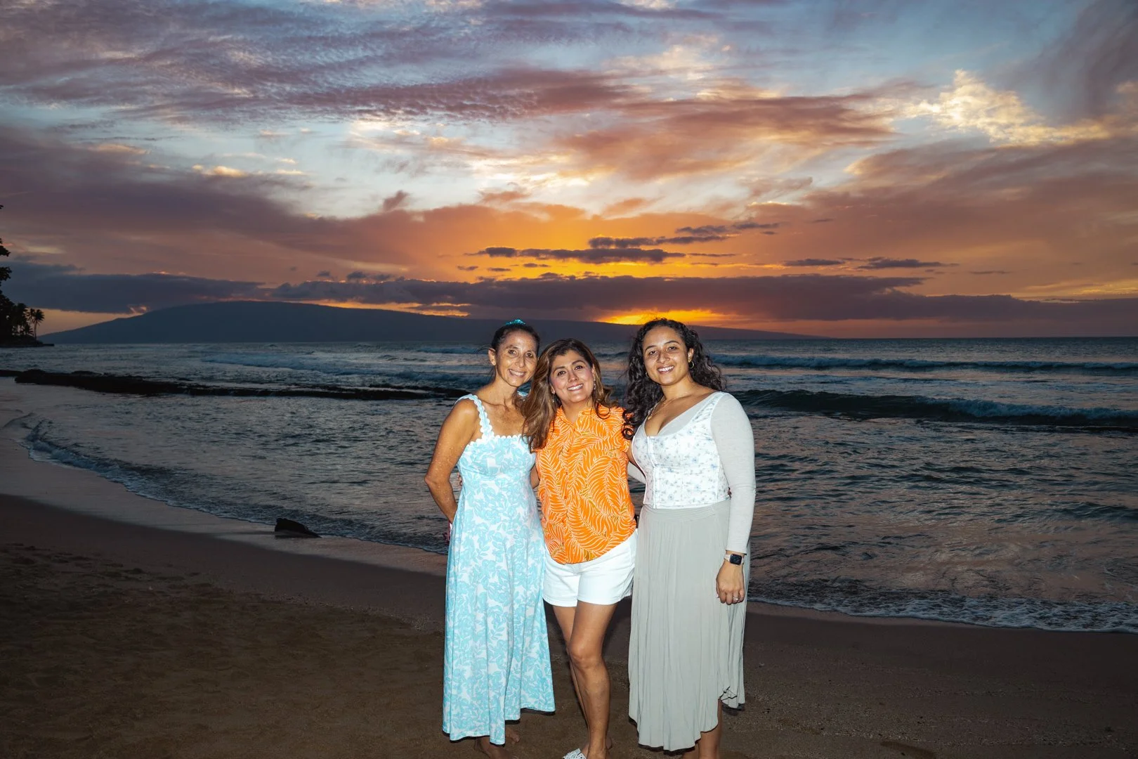 Three women smiling on the beach during sunset with the ocean and sky in the background.