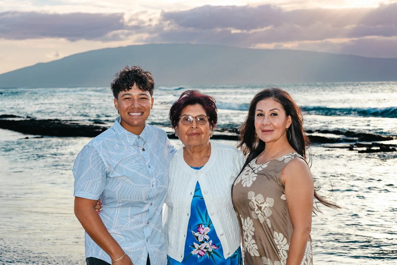 A family of three women and one young man posing on a beach at sunset, with ocean waves and a mountain in the background.