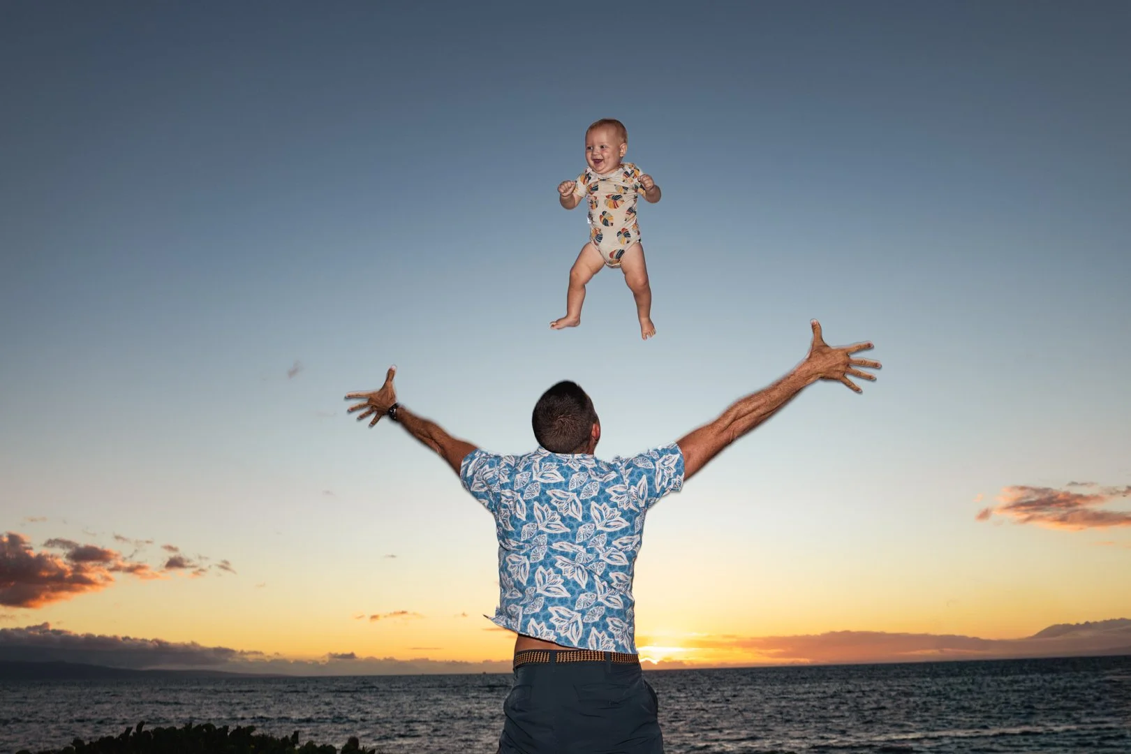 Man in a blue Hawaiian shirt lifting a smiling baby in a onesie into the air on a beach at sunset.