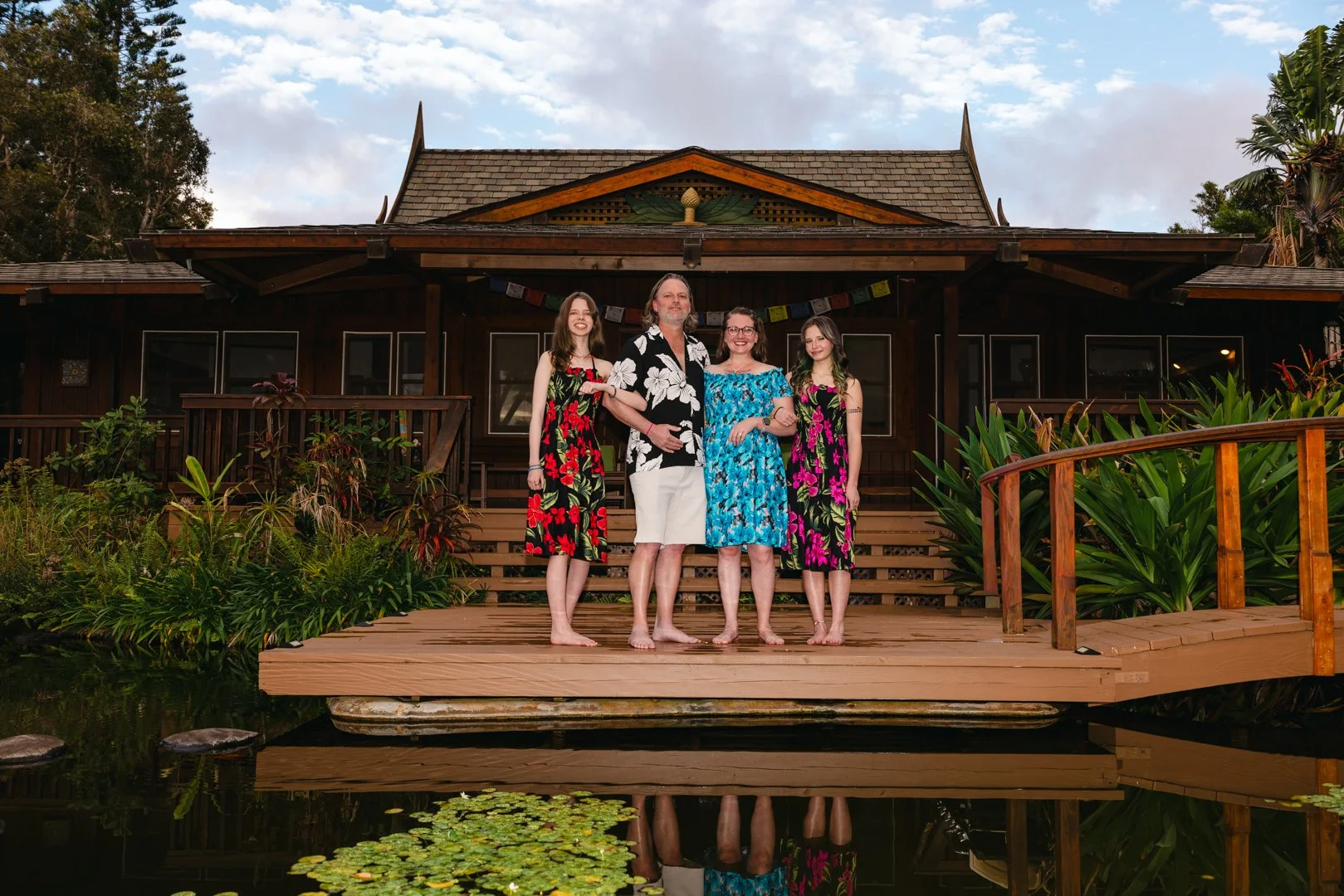 Four people standing on a wooden dock in front of a dark wooden house surrounded by lush greenery and a pond with floating lily pads.