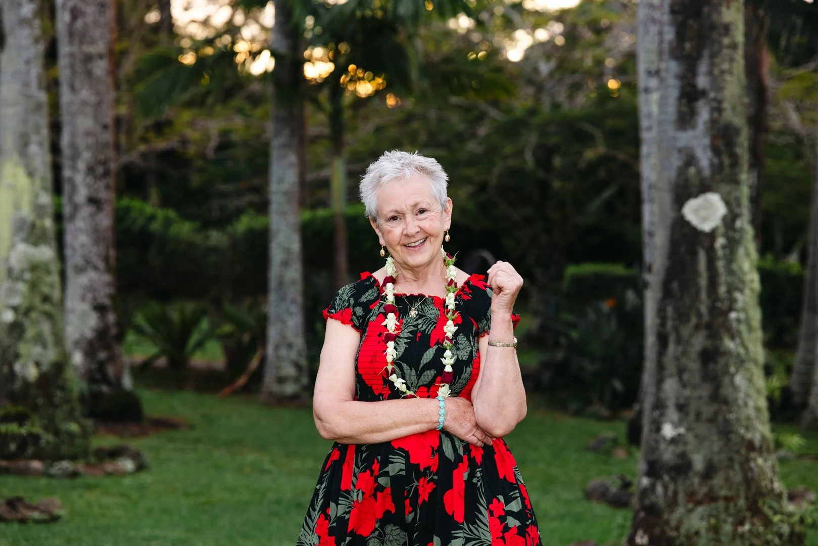 An elderly woman with short white hair standing outdoors with trees in the background, smiling, wearing a black and red floral dress, a flower lei, jewelry, and a bracelet.