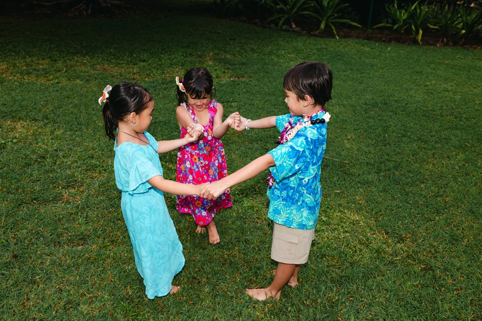 Three children, two girls and one boy, are holding hands and playing together on a grassy field. They are smiling and wearing colorful island-themed clothing.