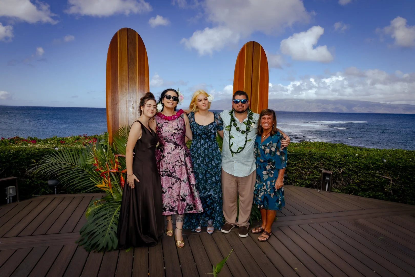 A group of five people standing on a wooden deck with the ocean and sky in the background, dressed in tropical clothing and leis, posed in front of two large wooden surfboard decorations.