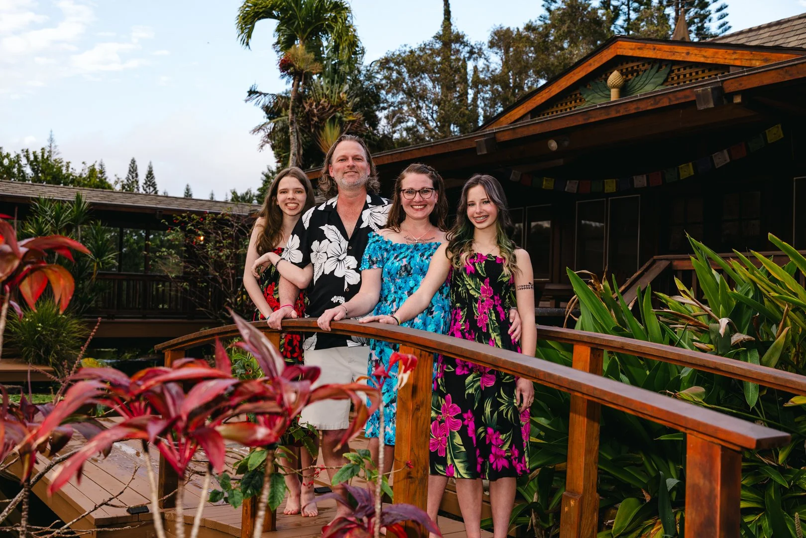 A family of four standing on a wooden bridge outdoors, surrounded by lush tropical plants and trees, with a house in the background, all dressed in colorful, floral-patterned clothing.