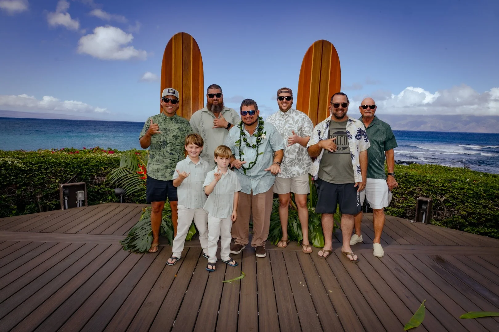 A group of seven men and two children standing on a wooden deck near the ocean with surfboards in the background, wearing tropical shirts, sunglasses, and casual summer attire.