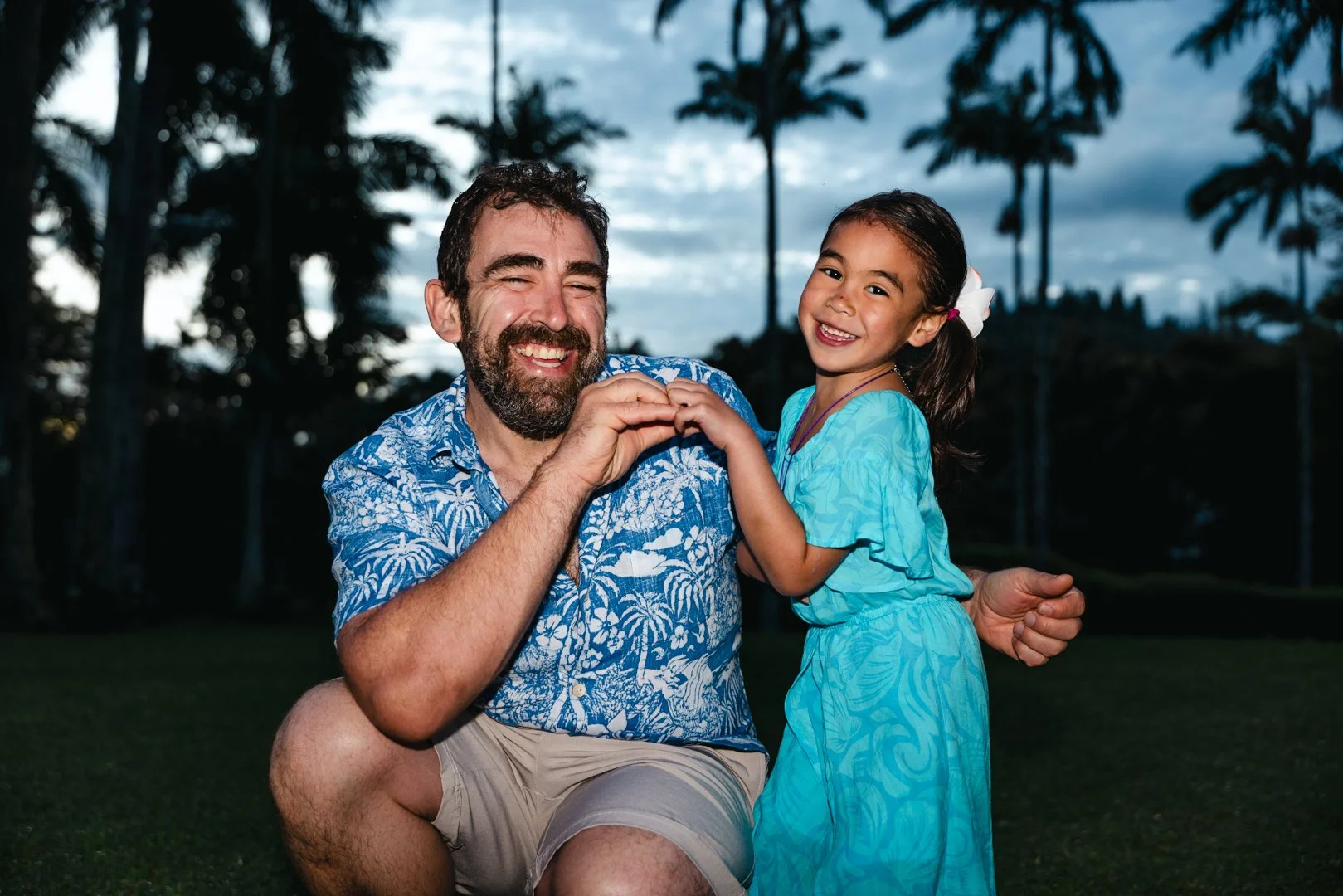A smiling man and young girl making a heart shape with their hands outdoors at sunset, with palm trees in the background