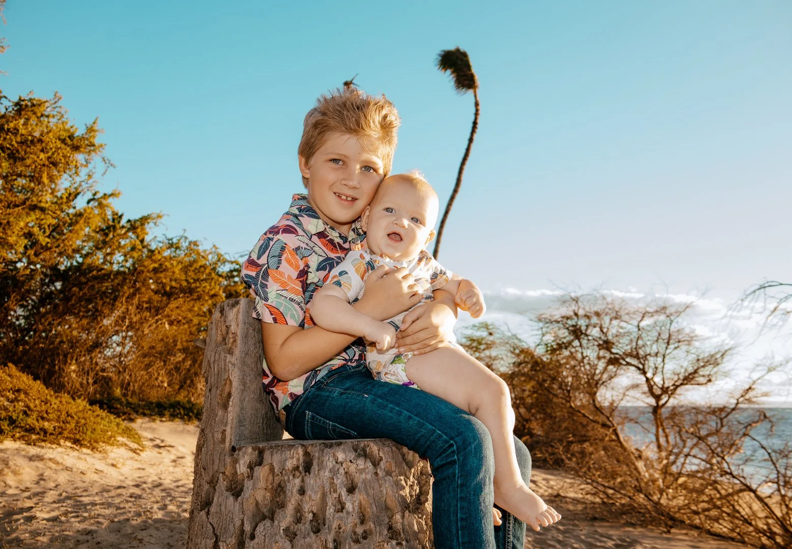 Two children, a boy and a baby girl, sitting on a tree stump at the beach with trees and a clear blue sky in the background.