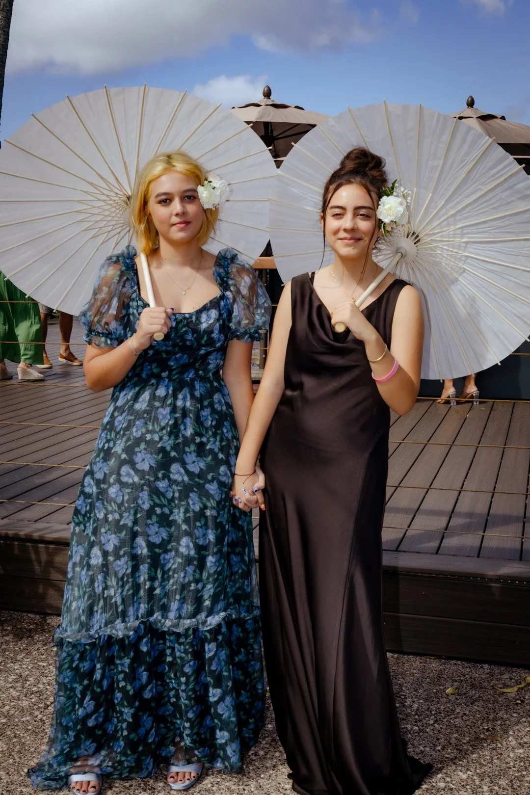 Two young women standing side-by-side on a wooden deck, holding white parasols adorned with flowers. They are dressed in elegant, long dresses and are holding hands. The background features brown umbrellas and a partly cloudy sky.