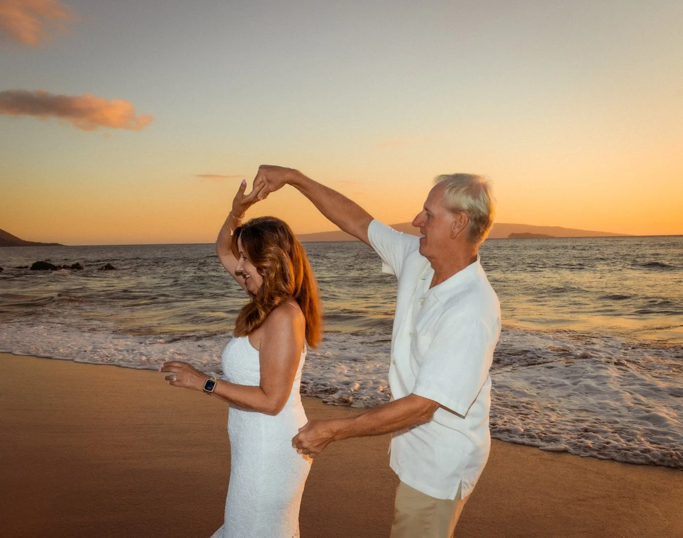 An older man and a woman dance on a beach at sunset, the man twirling the woman as they smile and enjoy the moment.