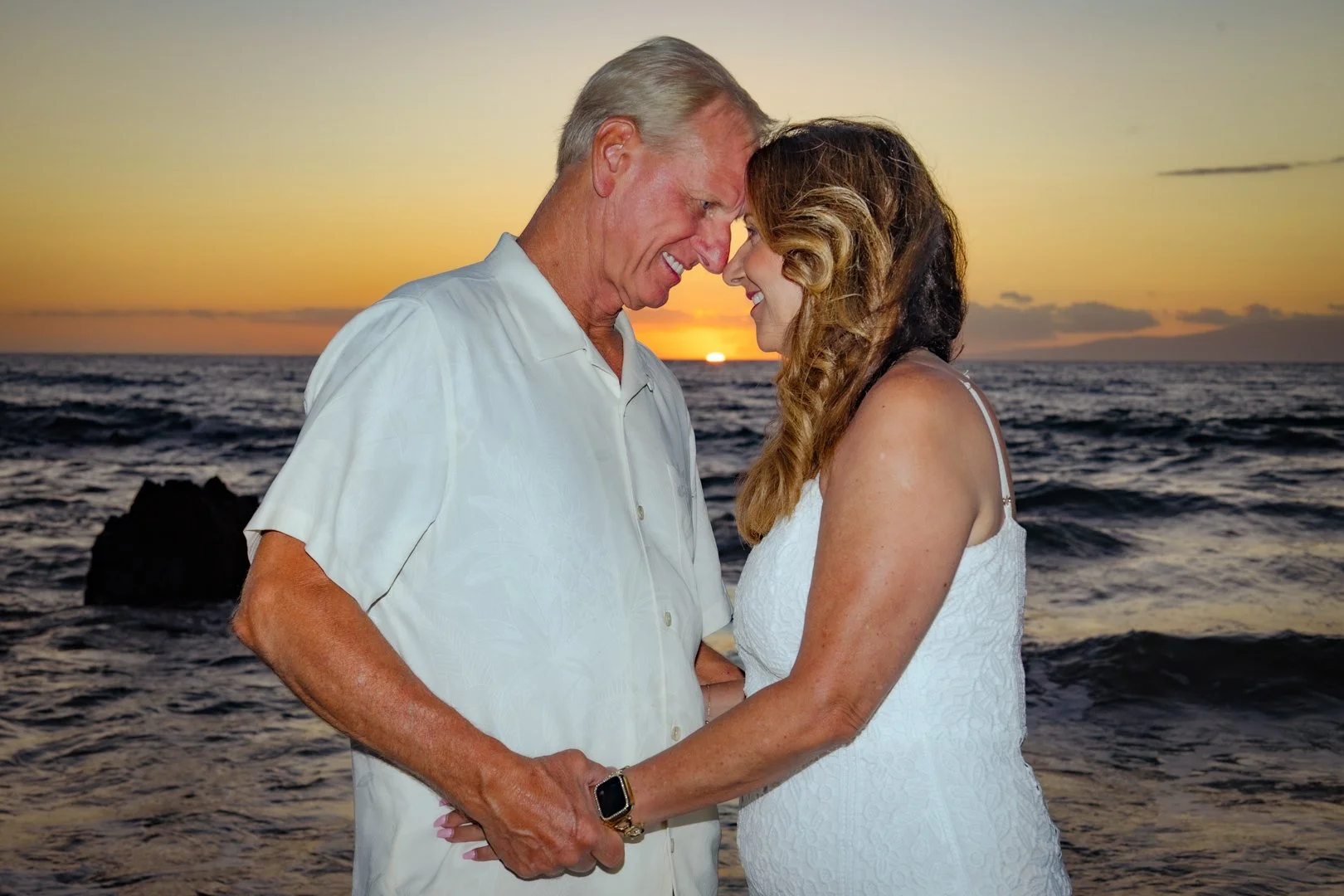An elderly man and a woman with long hair, both smiling, standing close together on the beach at sunset, with the ocean behind them.