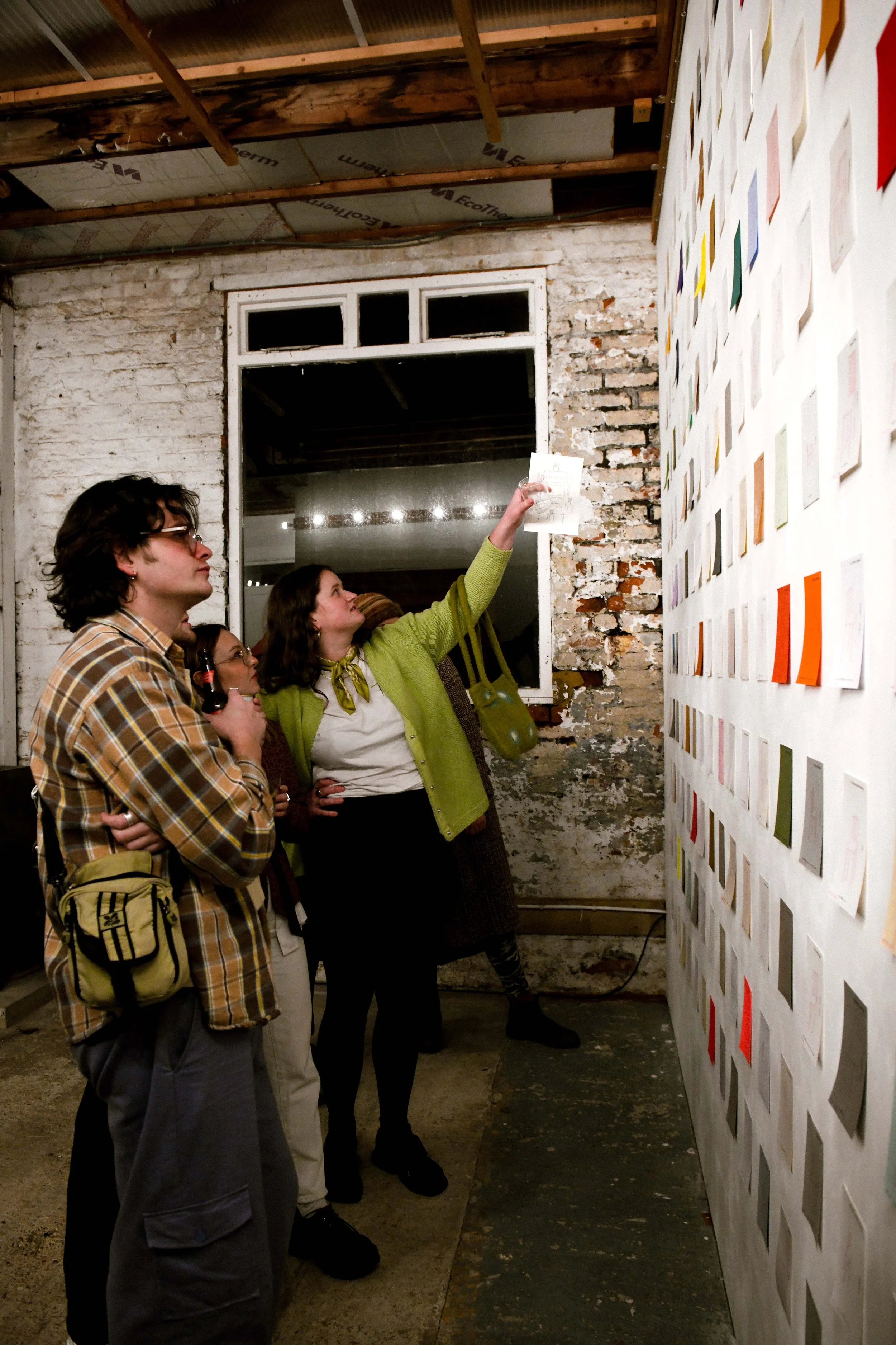 A small group of people stand in a stripped‑back, industrial room, looking over a wall covered in bright, handwritten notes. Someone points out a detail and the others lean in.