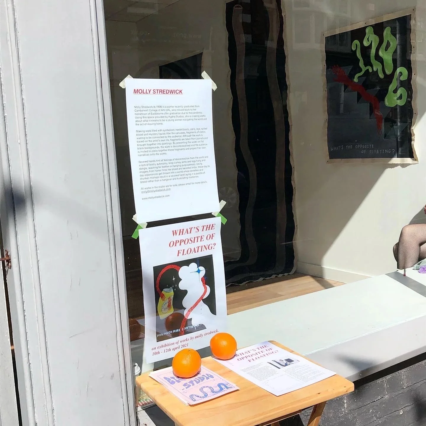 Exterior view of an exhibition space with posters and printed text displayed outside, alongside a table with handouts and oranges arranged for visitors.