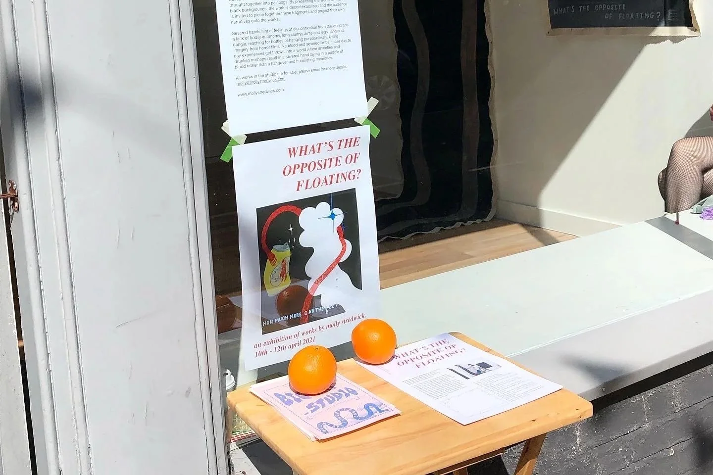 Photograph of Molly Stredwick's exhibition window in Eastbourne. The show was called 'What's The Opposite of Floating?' The window shows posters, small printed pieces and abstract artworks inside the gallery.
