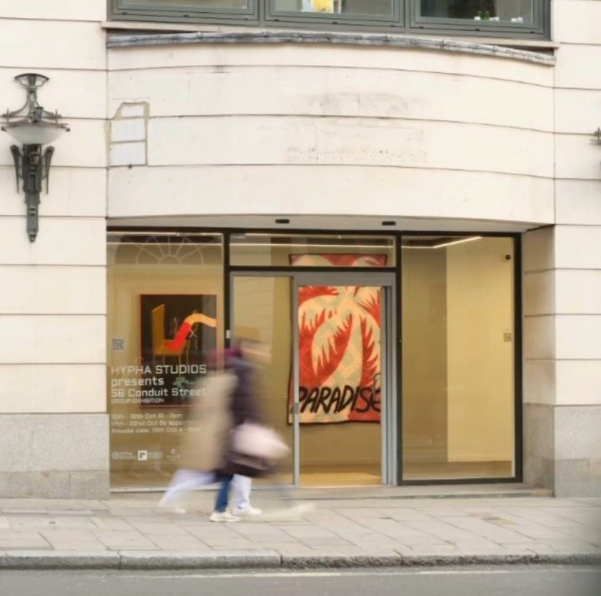 The exterior of a contemporary gallery on Conduit Street, with a bold red‑and‑orange artwork reading “PARADISE” visible through the glass entrance. Exhibition details are printed on the door, and two people walk past in motion blur.