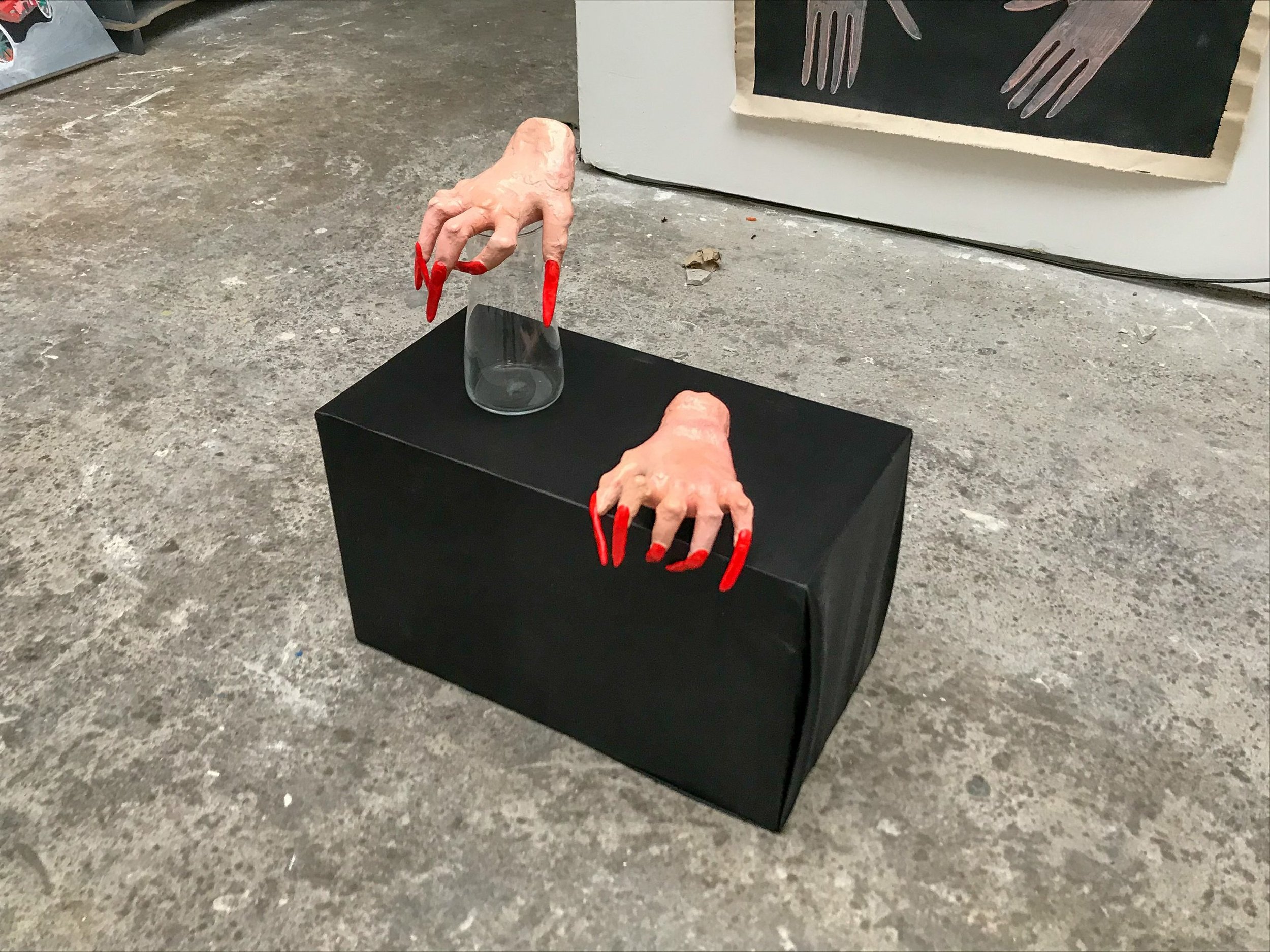 A sculptural installation featuring two lifelike hands with exaggerated red nails placed on a black plinth, one propped on an upside‑down glass. A painted hand study sits behind the piece, all set on a concrete floor in a minimal studio‑style space.