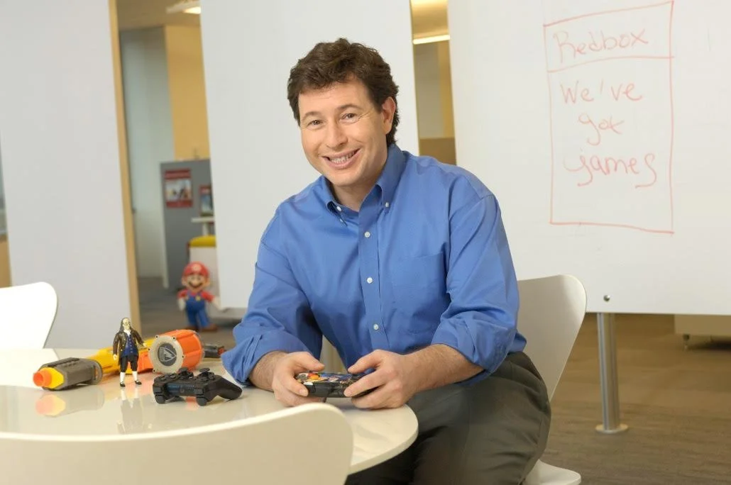 Mark Achler sitting at a white table with video game controllers, a Luigi figurine, and toy tools. In the background, there is a whiteboard with red writing and a Mario character figure.