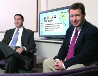 Two men sitting in an office, smiling, with a presentation screen in the background displaying text and graphics.