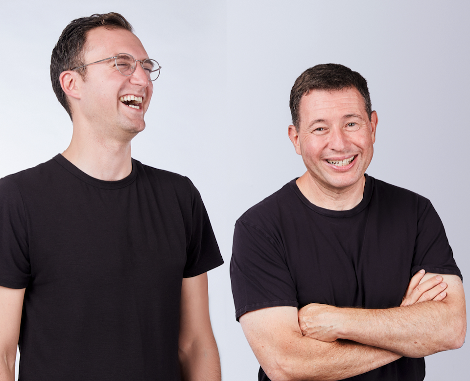 Two men wearing black t-shirts laughing and smiling, standing against a plain light background.