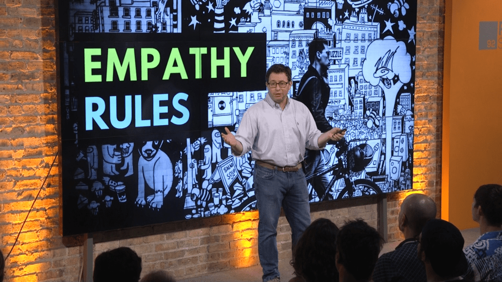 Mark Achler on stage, delivering a presentation to an audience about empathy rules, with a black-and-white illustrated cityscape on a large screen behind him.