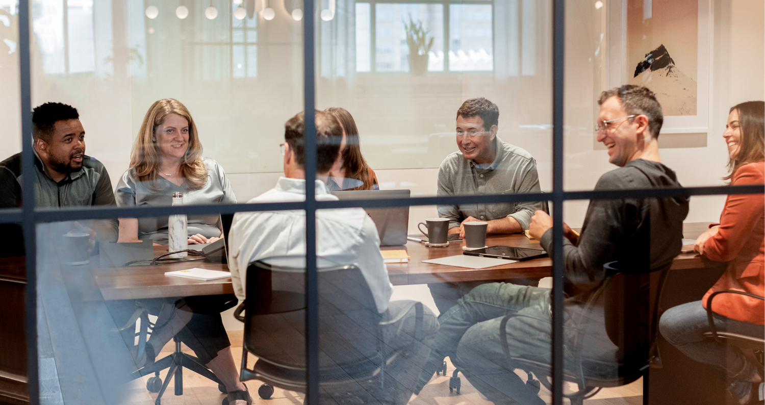 Group of seven diverse people sitting around a conference table having a discussion, visible through glass walls.