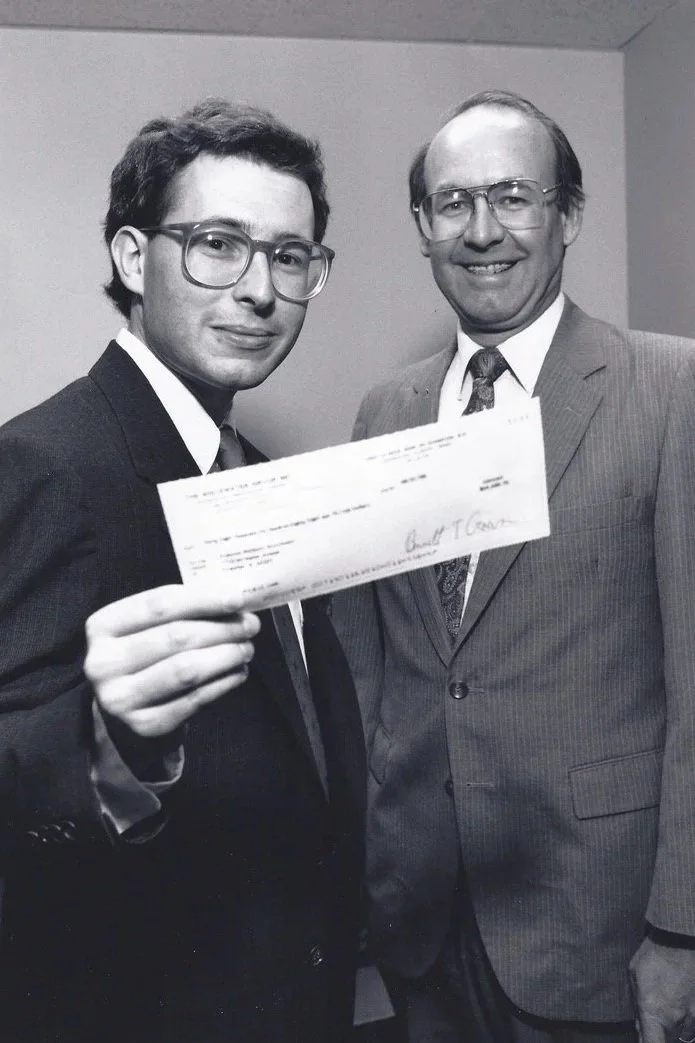 Two men in suits and glasses, one younger and one older, smiling and holding a check together in a black-and-white photo.