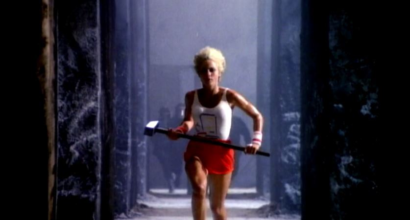 Woman with blond hair wearing a white tank top and red shorts holding a sledgehammer walking through a dark corridor.