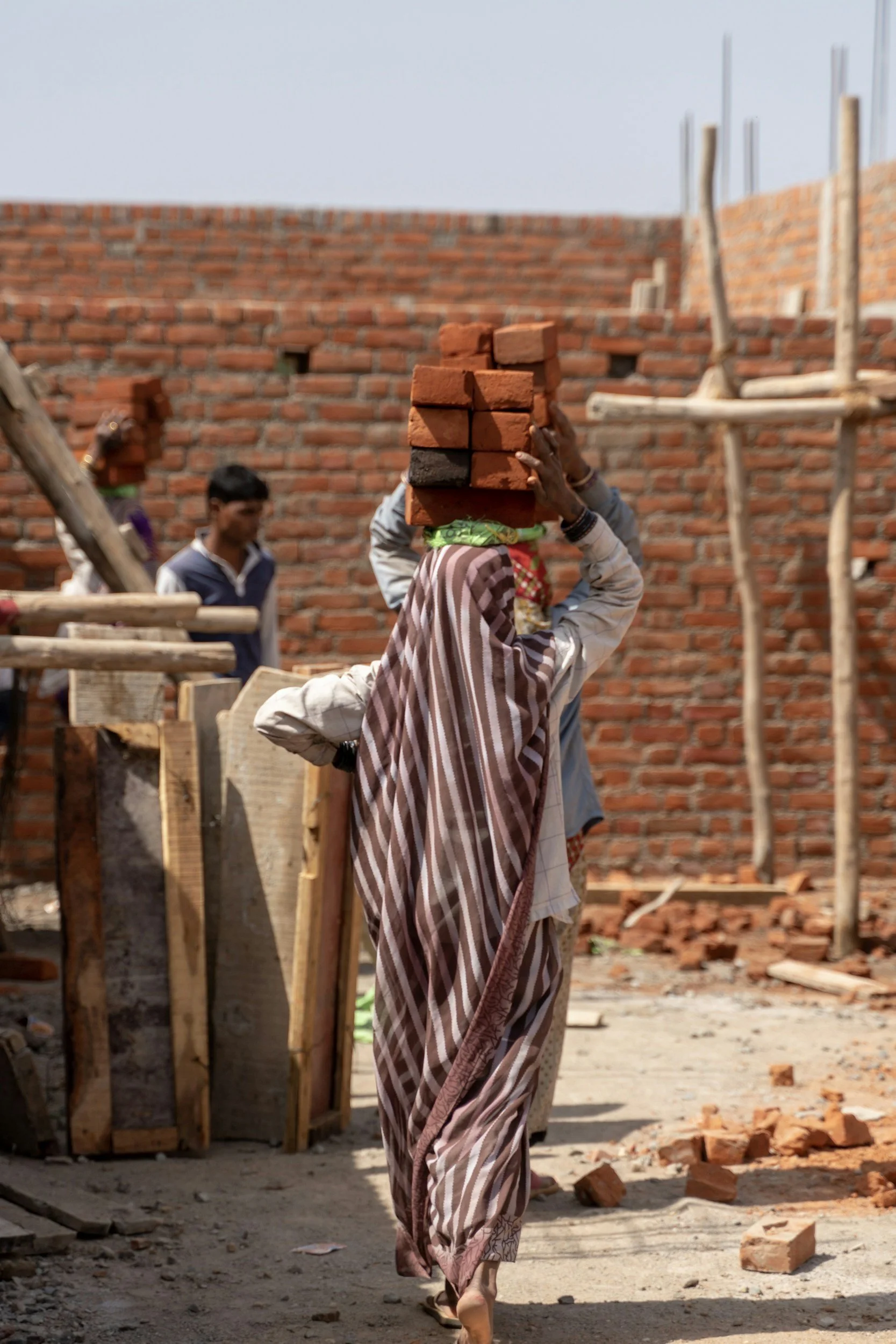 A woman carrying a stack of bricks on her head at a construction site with two men working and a partially built brick wall in the background.