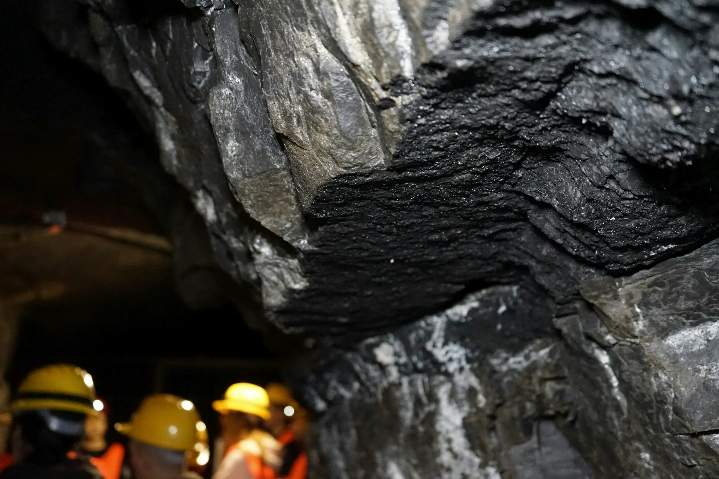 Mining cave with dark, jagged rock formations and a group of miners wearing yellow safety helmets and orange safety vests in the background.