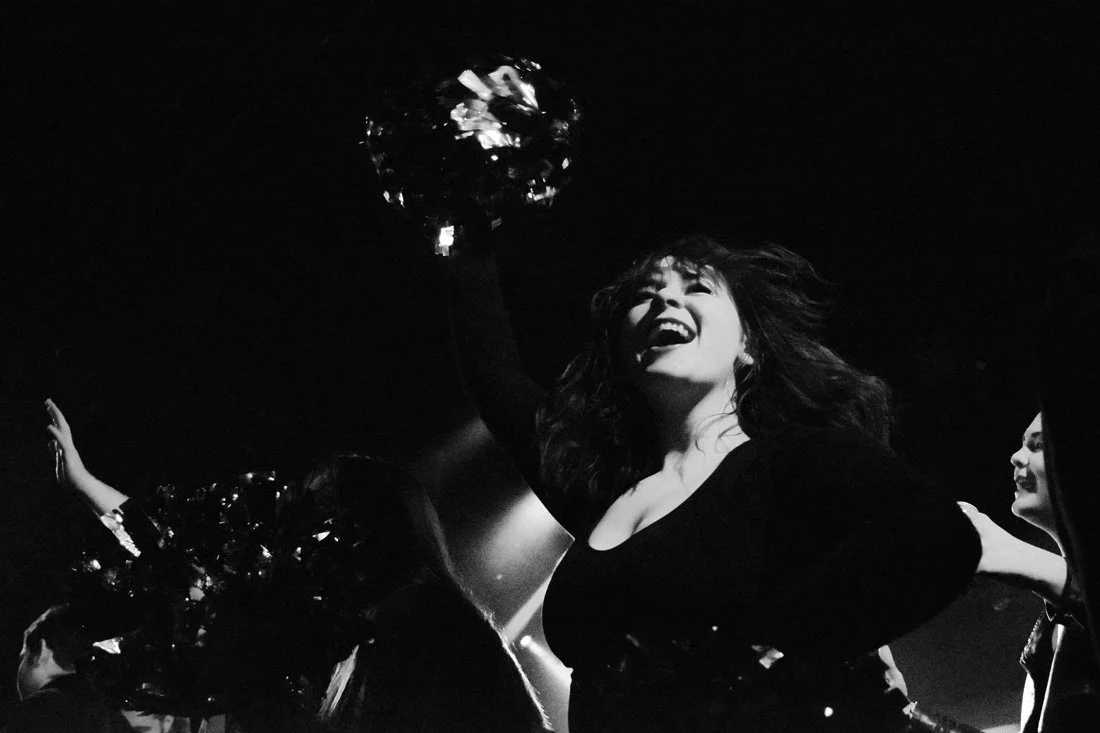 A woman cheerleader holding pom-poms, smiling with her hair flying, in a dark setting with other cheerleaders.