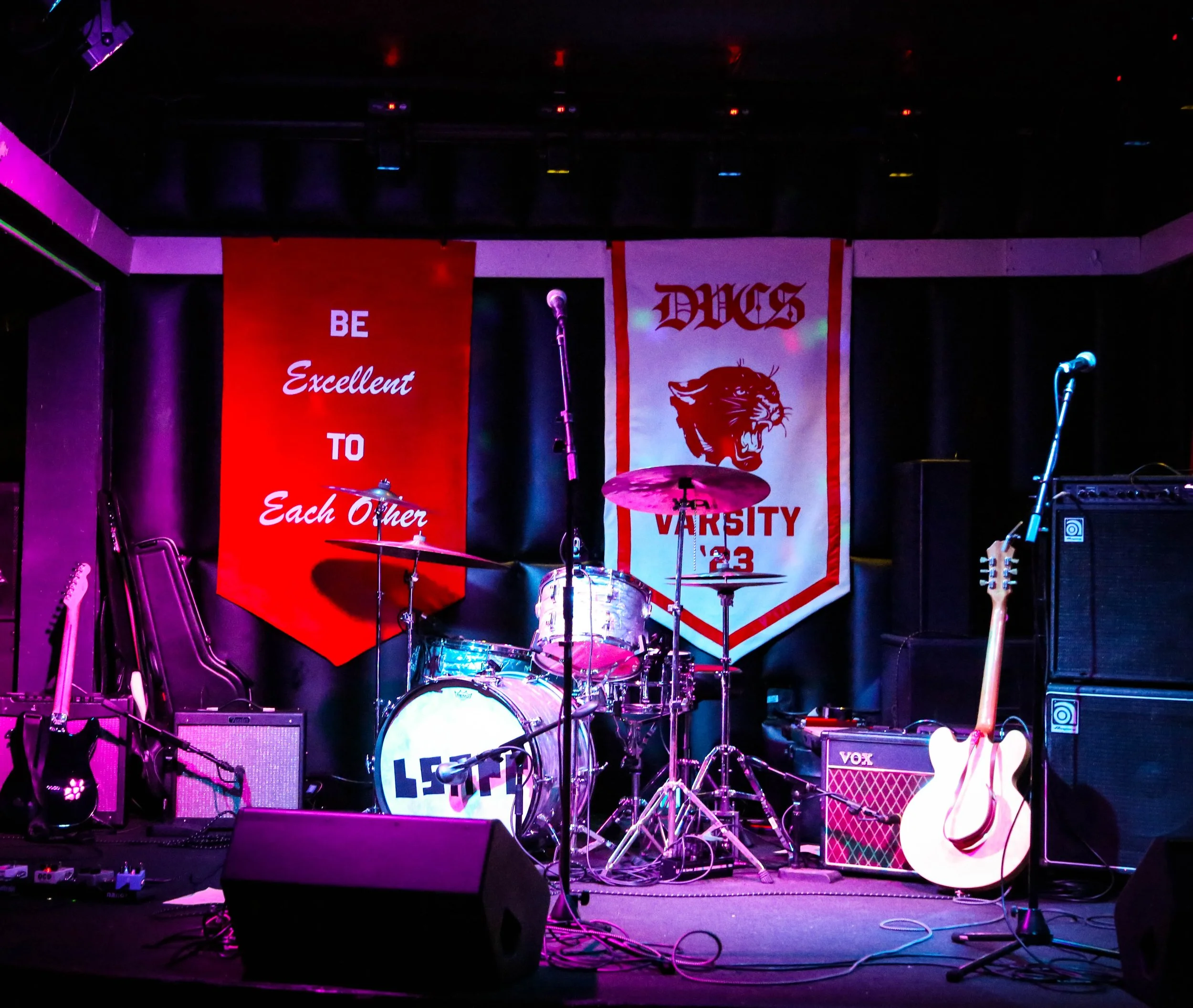 A stage setup with musical instruments including drums, guitars, microphones, and amplifiers. Behind the stage, there are two banners, one red with the text 'BE Excellent TO Each Other' and the other a school or team banner with a tiger logo. The stage is lit with purple lighting and the scene appears to be prepared for a performance or event.