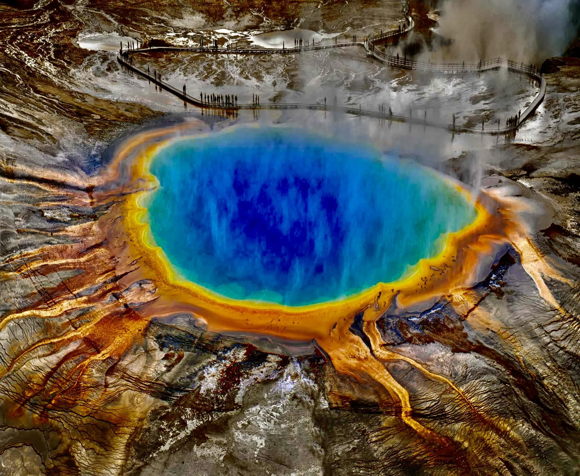 Colorful geothermal hot spring with vibrant blue center, surrounded by yellow, orange, and brown mineral deposits, steam rising in the background.