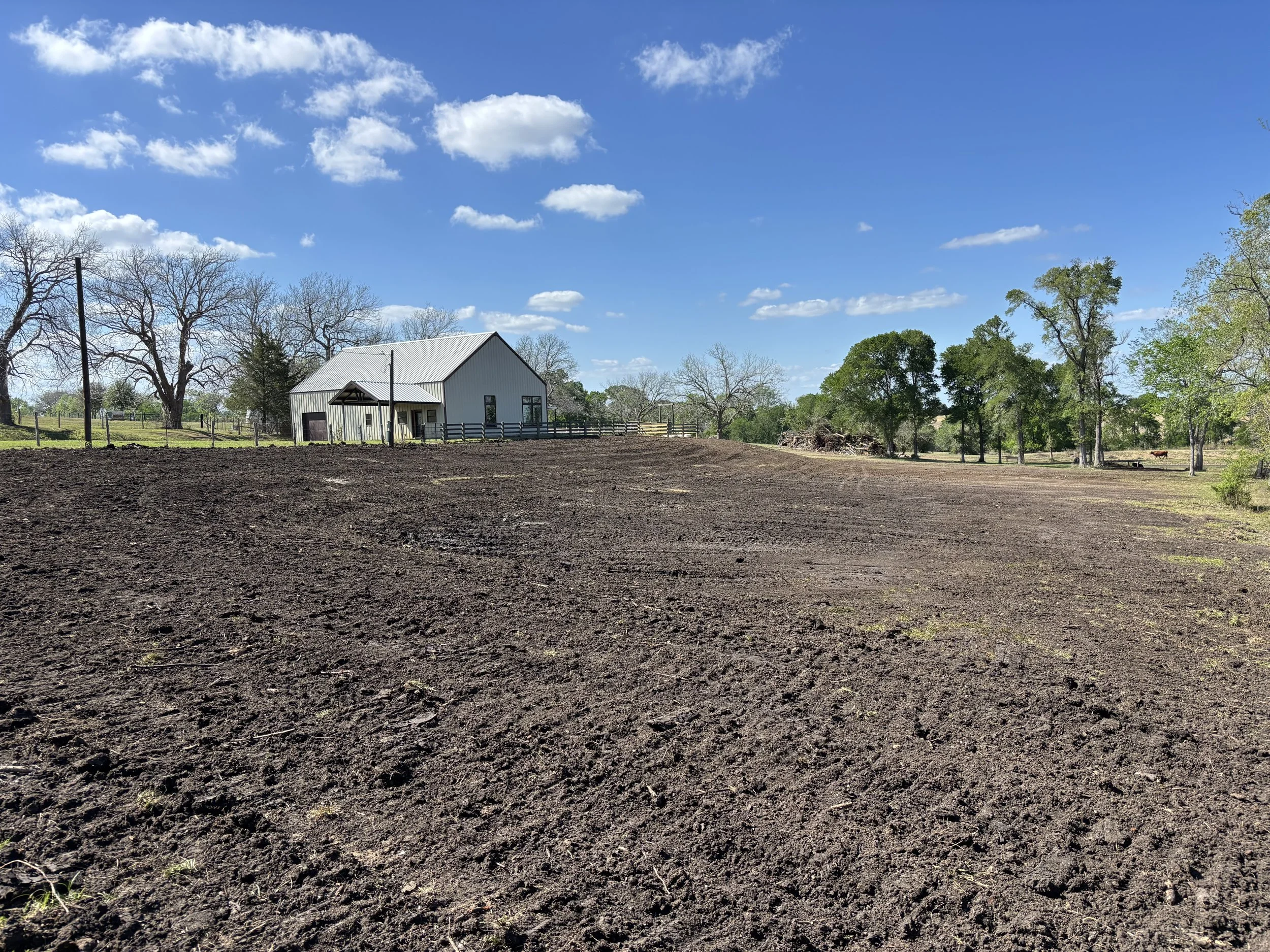 A white barn with a gray roof in a rural landscape under a blue sky with scattered clouds, surrounded by trees and a fenced area, with freshly tilled dark soil in the foreground.