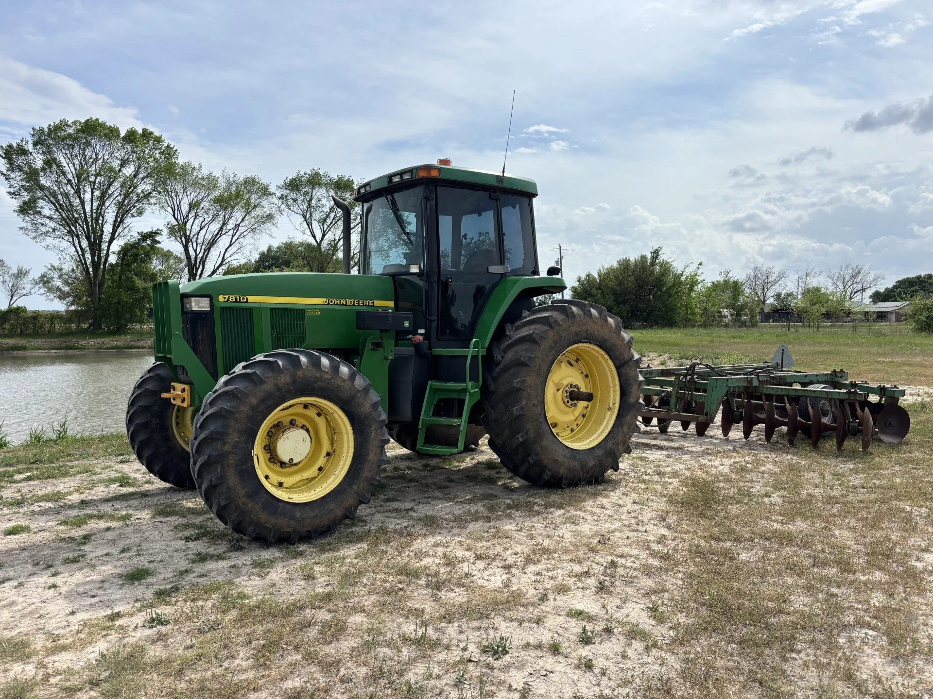 Green John Deere tractor with large black tires and a yellow wheel, attached to farming equipment, on a dirt area near a body of water with trees and houses in the background under a partly cloudy sky.