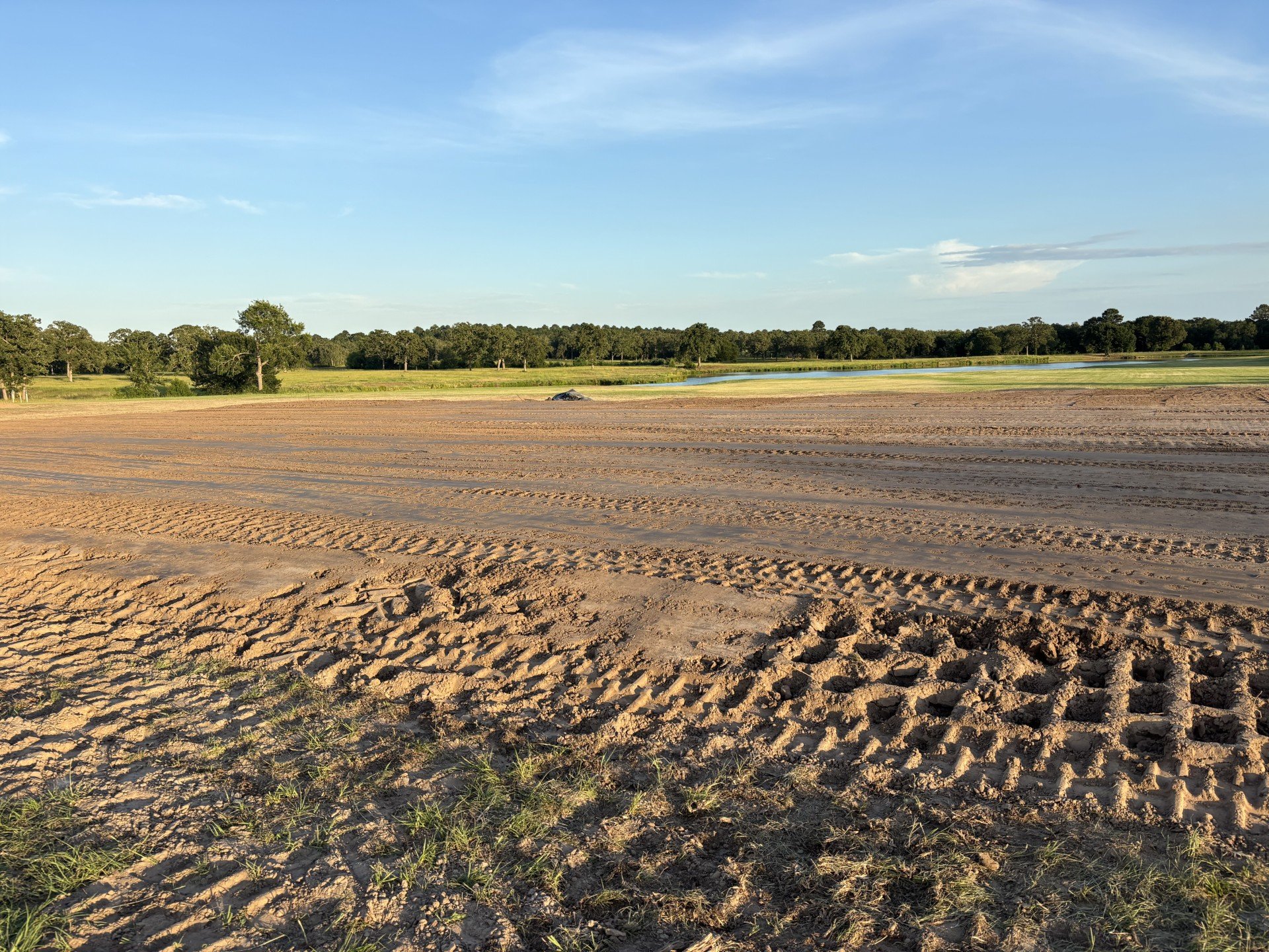 A dirt field with tire tracks in the foreground, green trees in the middle, and a lake and blue sky with clouds in the background.