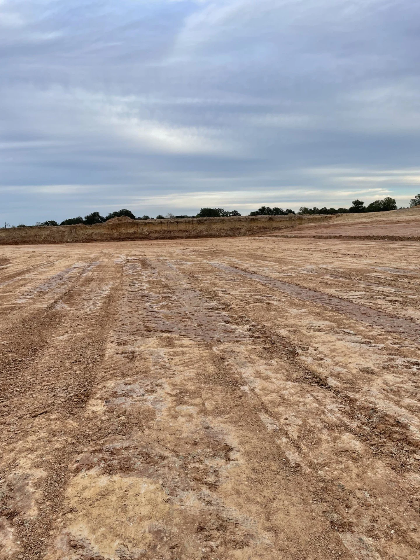 A wide, flat construction site with dirt and gravel, with a raised dirt embankment in the distance and a cloudy sky above.