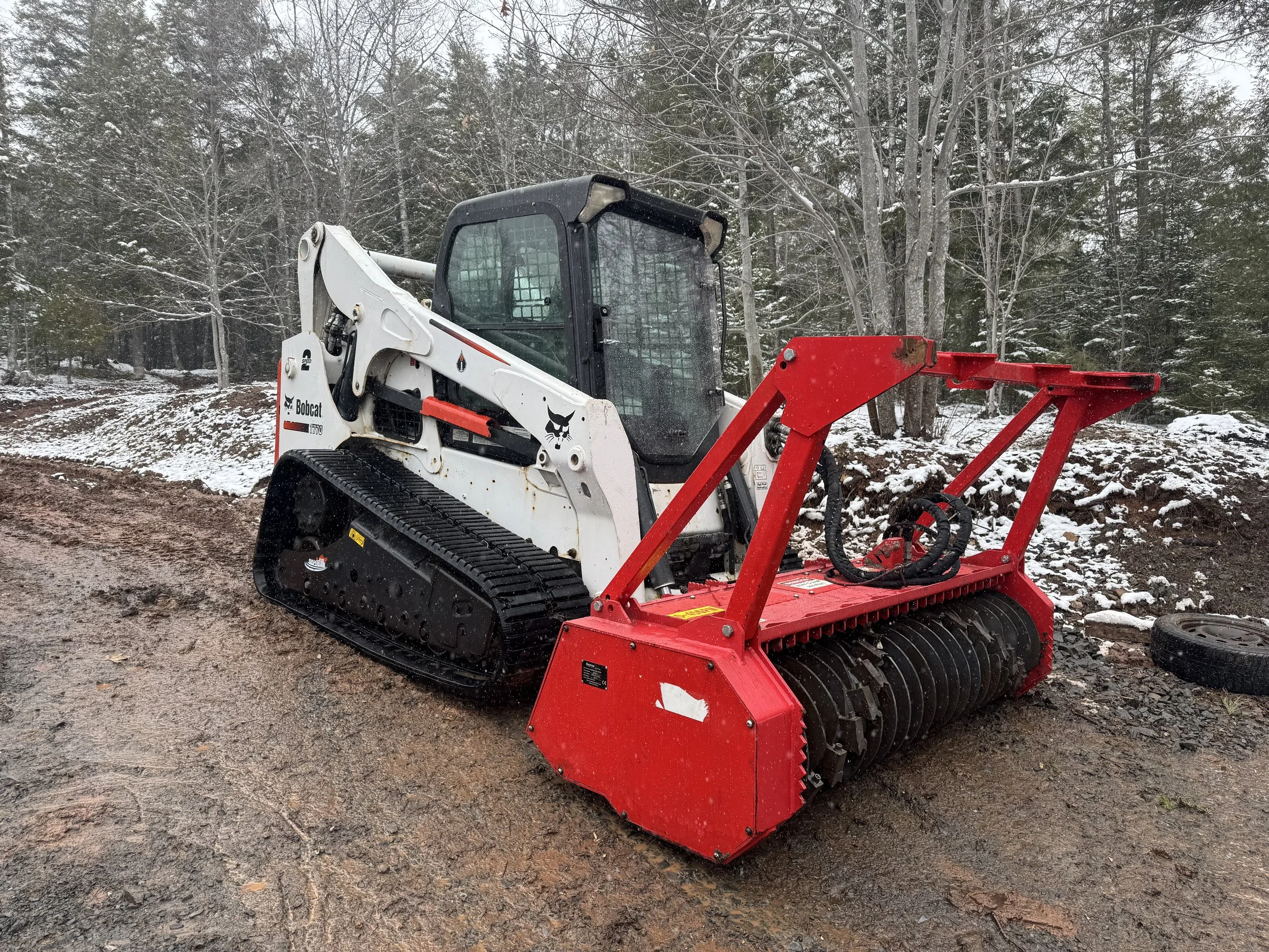 A Bobcat skid steer loader with mulcher attachment on a muddy, snow-dusted outdoor construction site, with trees in the background.