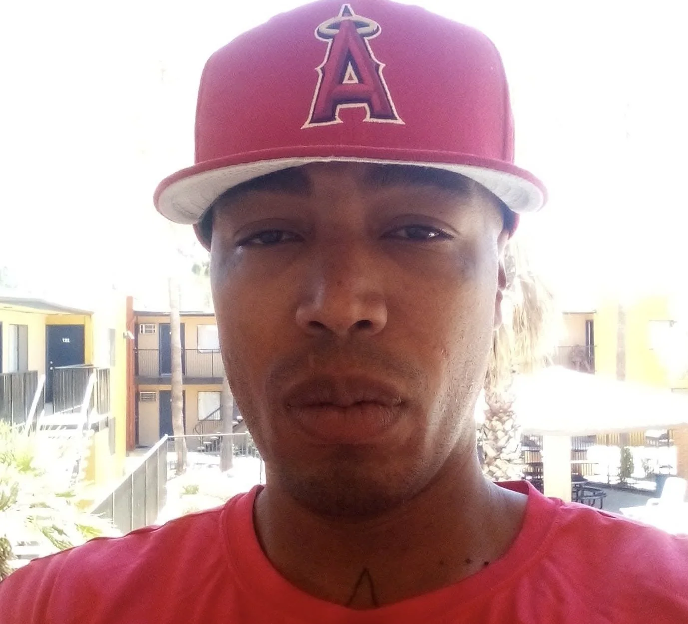 Close-up of a young man wearing a red baseball cap with the Los Angeles Angels logo and a red shirt, standing outdoors with apartment buildings and a palm tree in the background.
