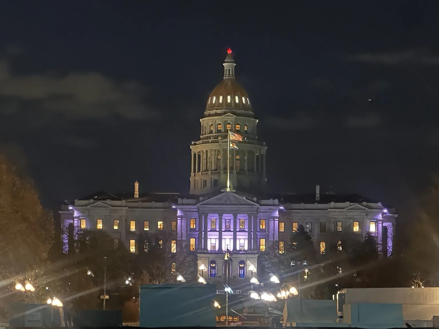 Nighttime view of Colorado State Capitol building illuminated with purple and white lights, with a flagpole and American flag in front.