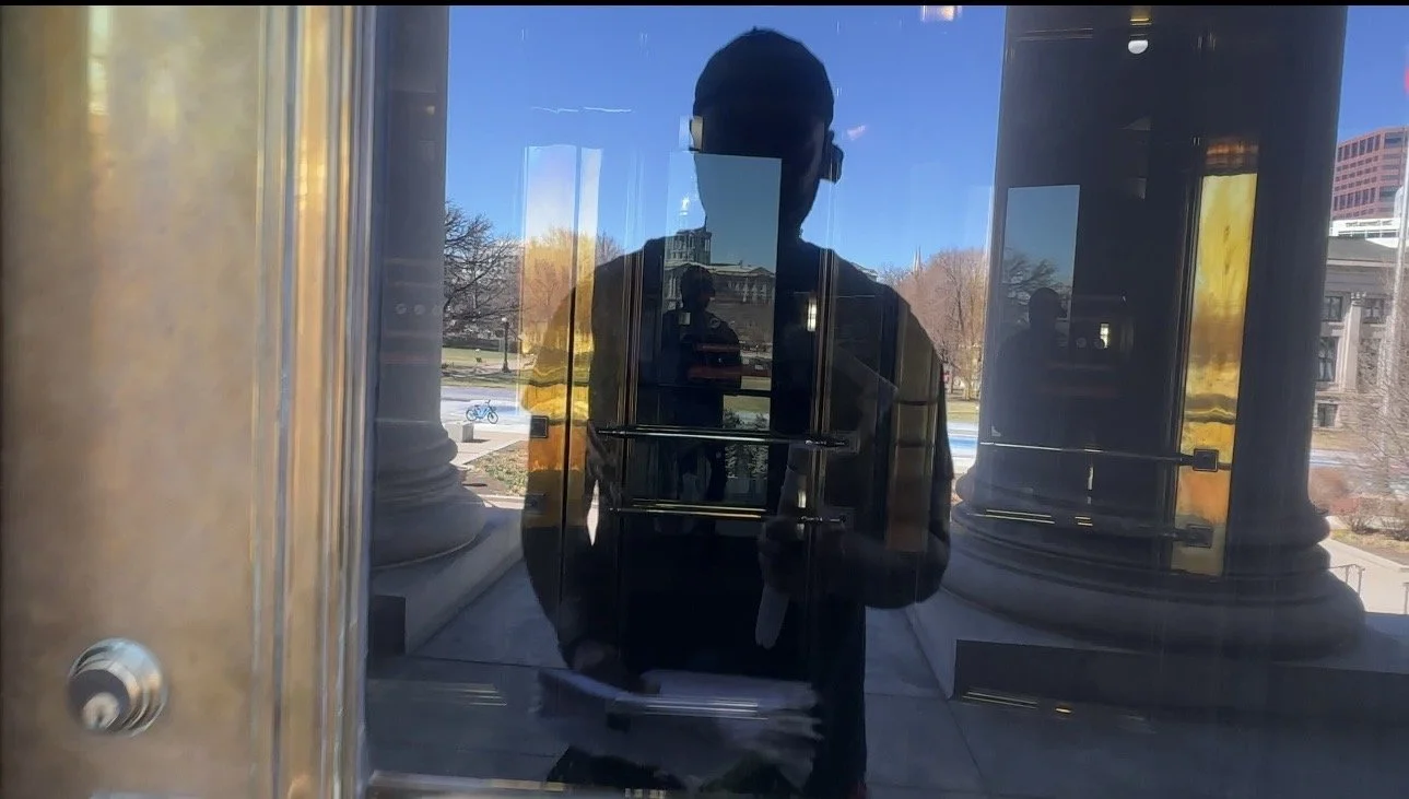 Reflection of Socrates Ulysses Packer taking a photo in a glass door or window outside of a building, with columns and trees visible in the background.